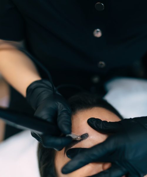 Partial image of beautician doing eyebrow microblading with needle tool of woman client in beauty salon. Semi-permanent makeup for eyebrows. Female master wearing gloves. Beauty concept